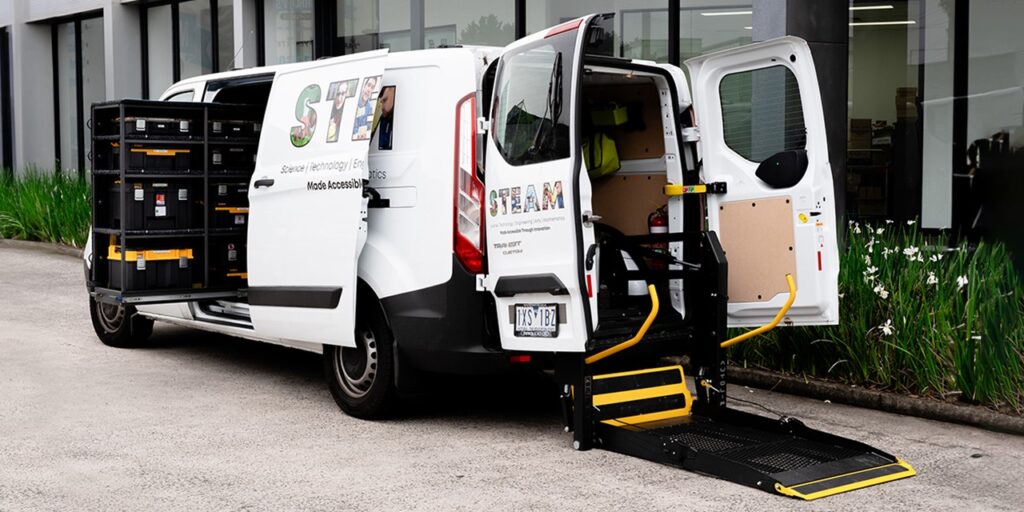 The inclusive Makervan. A white van with 'STEAM' written on the side and back, an equipment rack extending out the side door, and a wheelchair lift attached at the back.