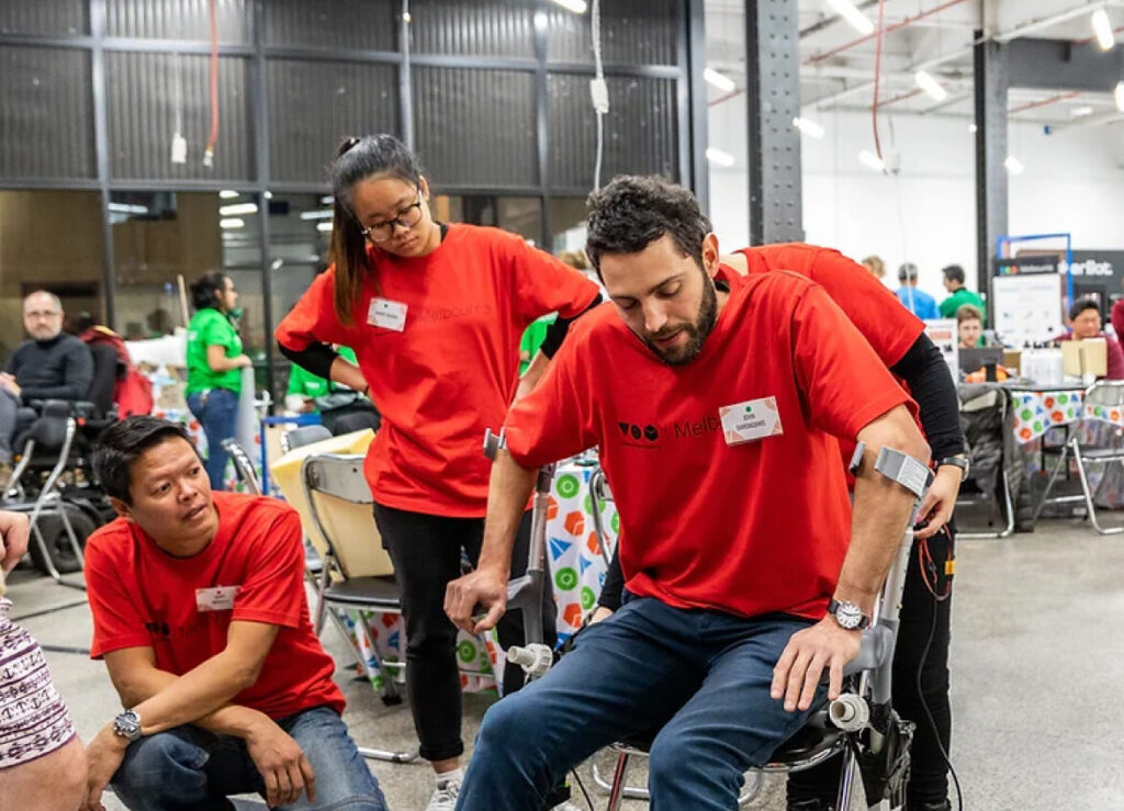 Two people in red shirts are watching while a third man in red is testing a prototype on a pair of crutches.