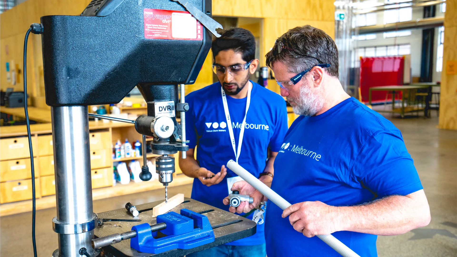 Two men in blue t-shirts are holding a pipe by a drill.