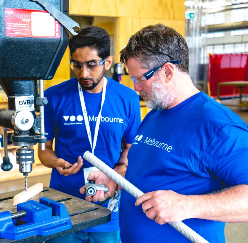 Two men in blue t-shirts are holding a pipe by a drill.