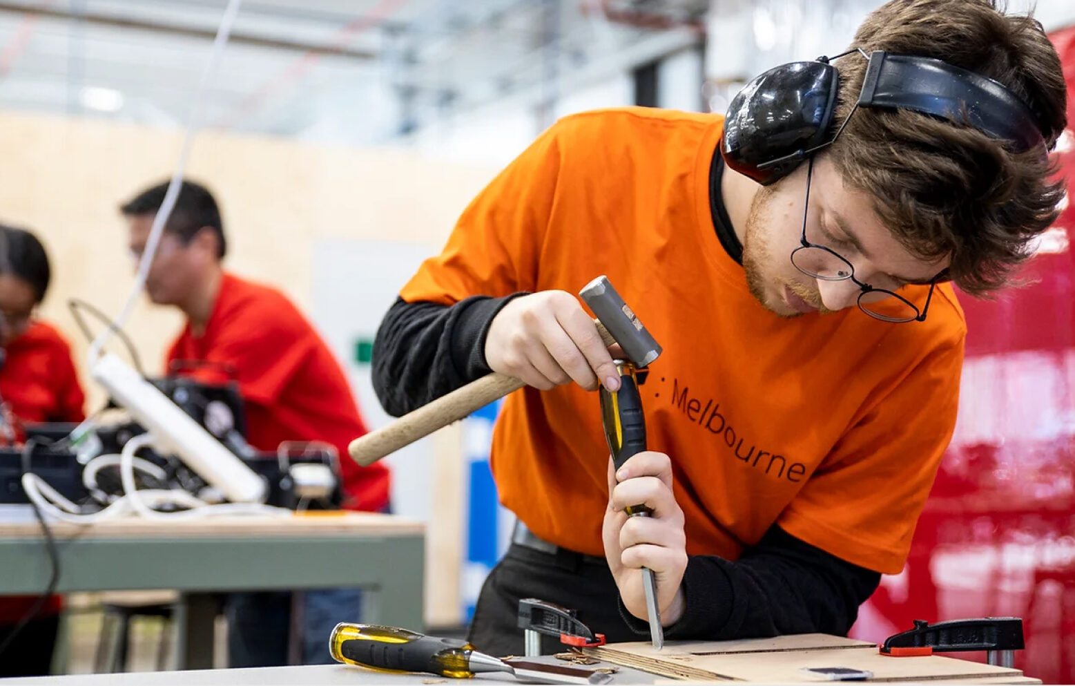 Man in orange TOM t-shirt using a hammer and chisel on a piece of plywood.