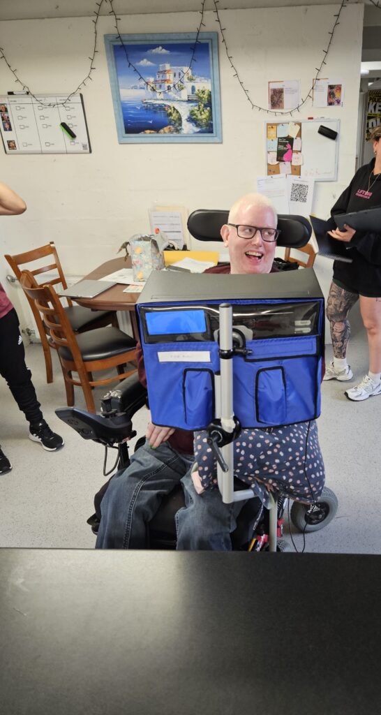 A man in a wheelchair is looking to the left and smiling behind his communication device.