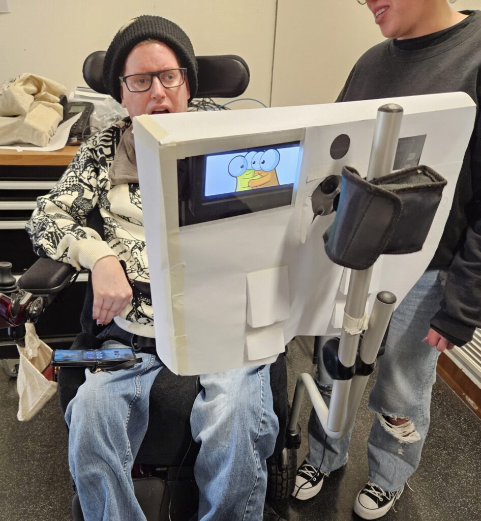 A man in a wheelchair is behind his communication device covered in a paper prototype.