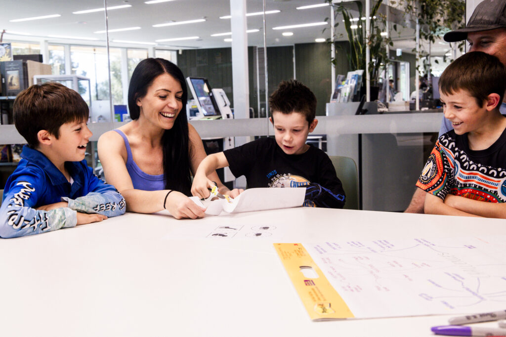 Young boy using scissors while his parents and two brothers are watching him and laughing.