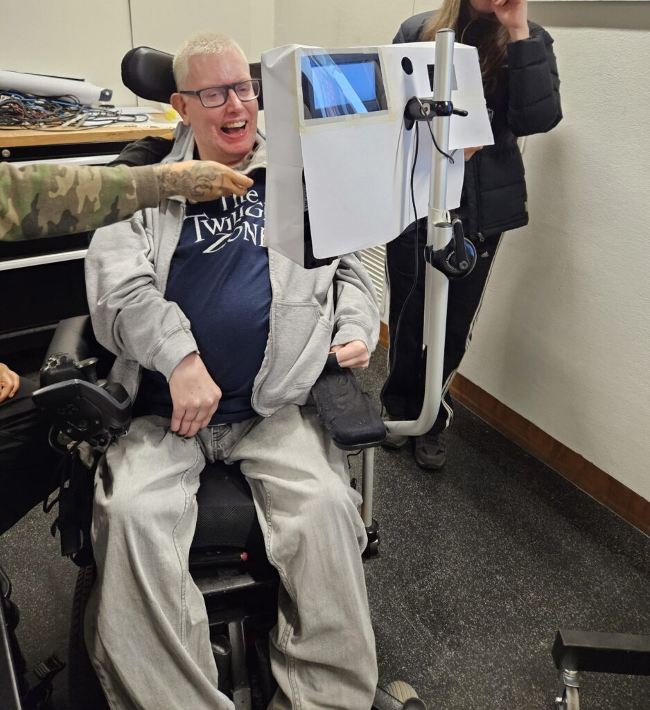A man in a wheelchair is smiling behind his communication device covered in a paper prototype.