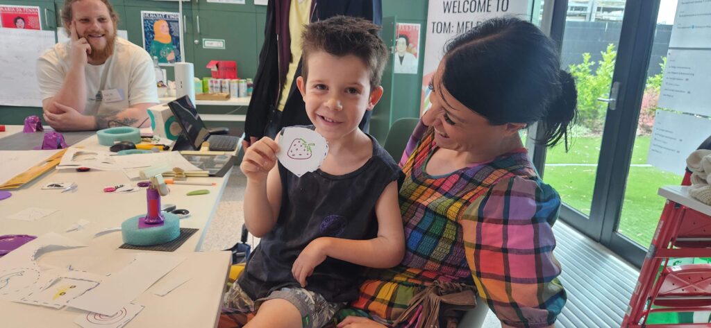 Boy is sitting on womans lap smiling holding a paper cut out of a strawberry.