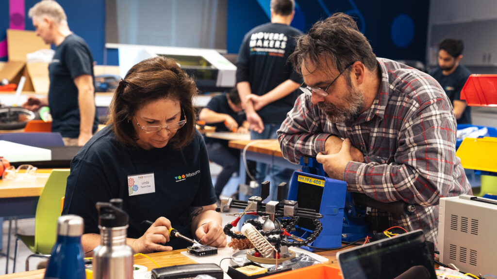 A man is watching a woman is soldering electronics.