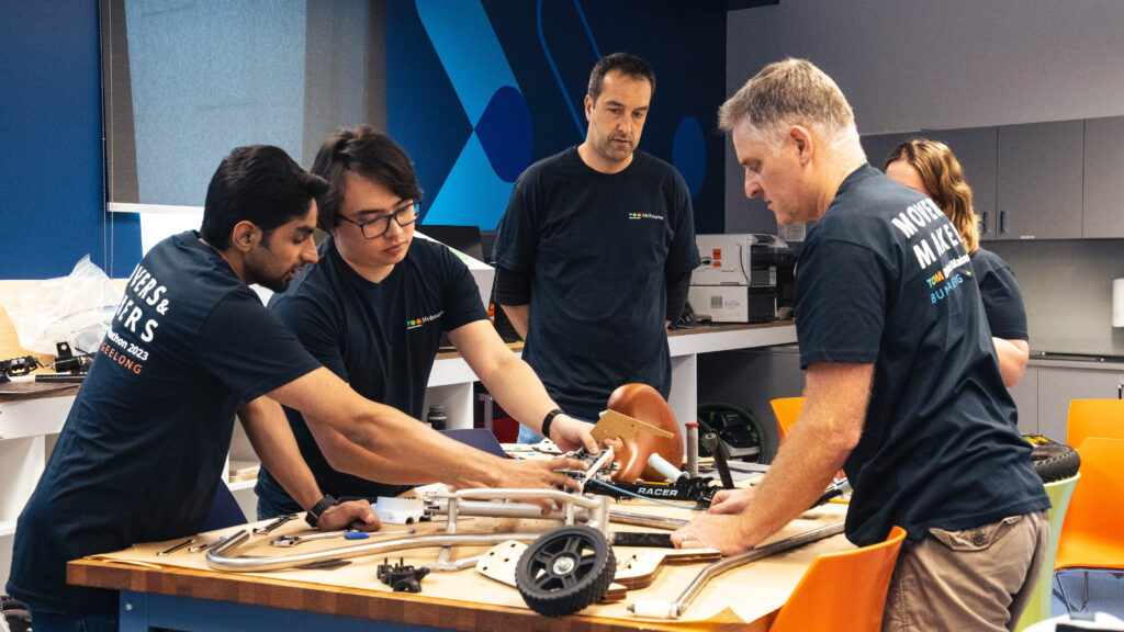 Five people in TOM t-shirts are working on a prototype by a work bench.