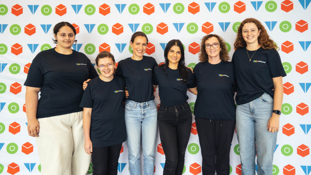 Six people in tom t-shirts are smiling to the camera in front of a tom picture wall.