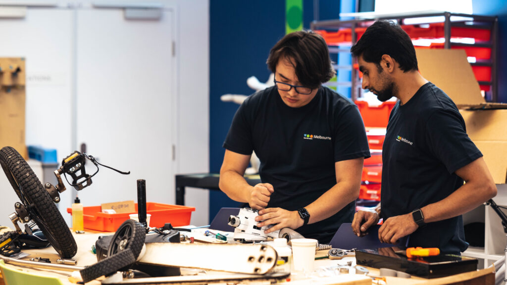Two people in TOM t-shirts are working on a prototype by a work bench.