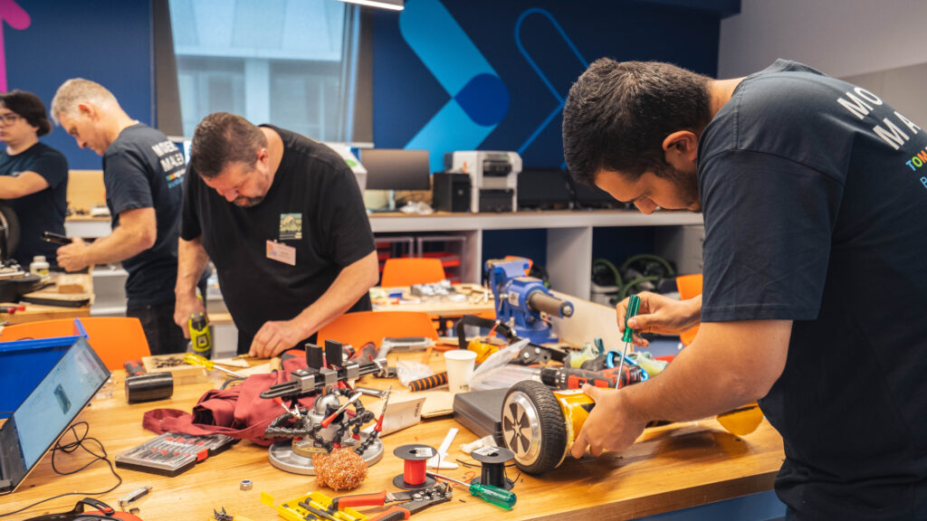 Two people drilling and screwing on a workbench covered with electronics.