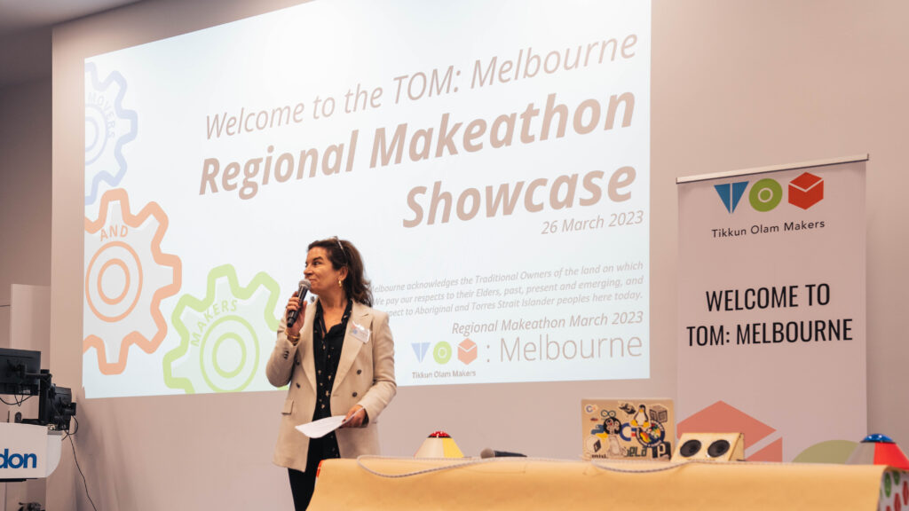 Woman on stage holding a microphone in front of a presentation saying "Regional Makeathon Showcase 26 March 2023".