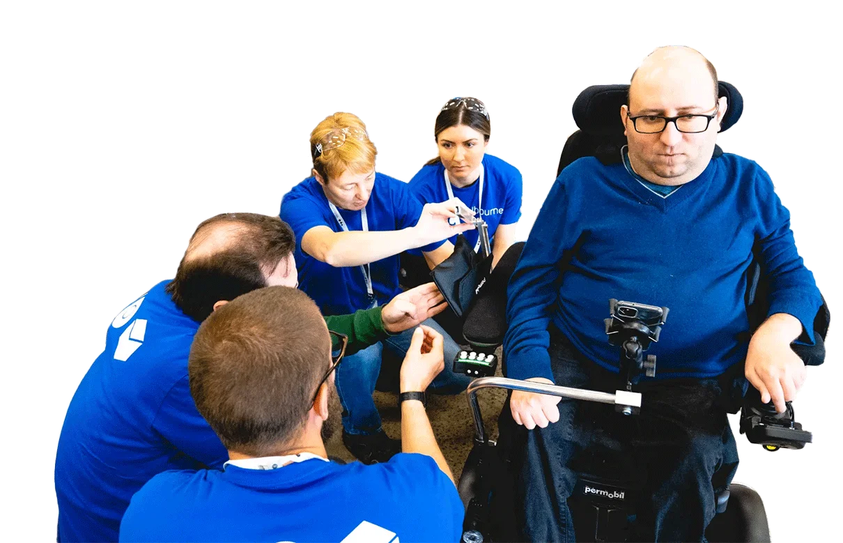 A man in a wheelchair is watching while four people in blue TOM t-shirts are working on a prototype attached to his wheelchair.