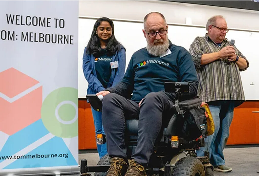 Three people on stage in front of a welcome to TOM Melbourne banner.